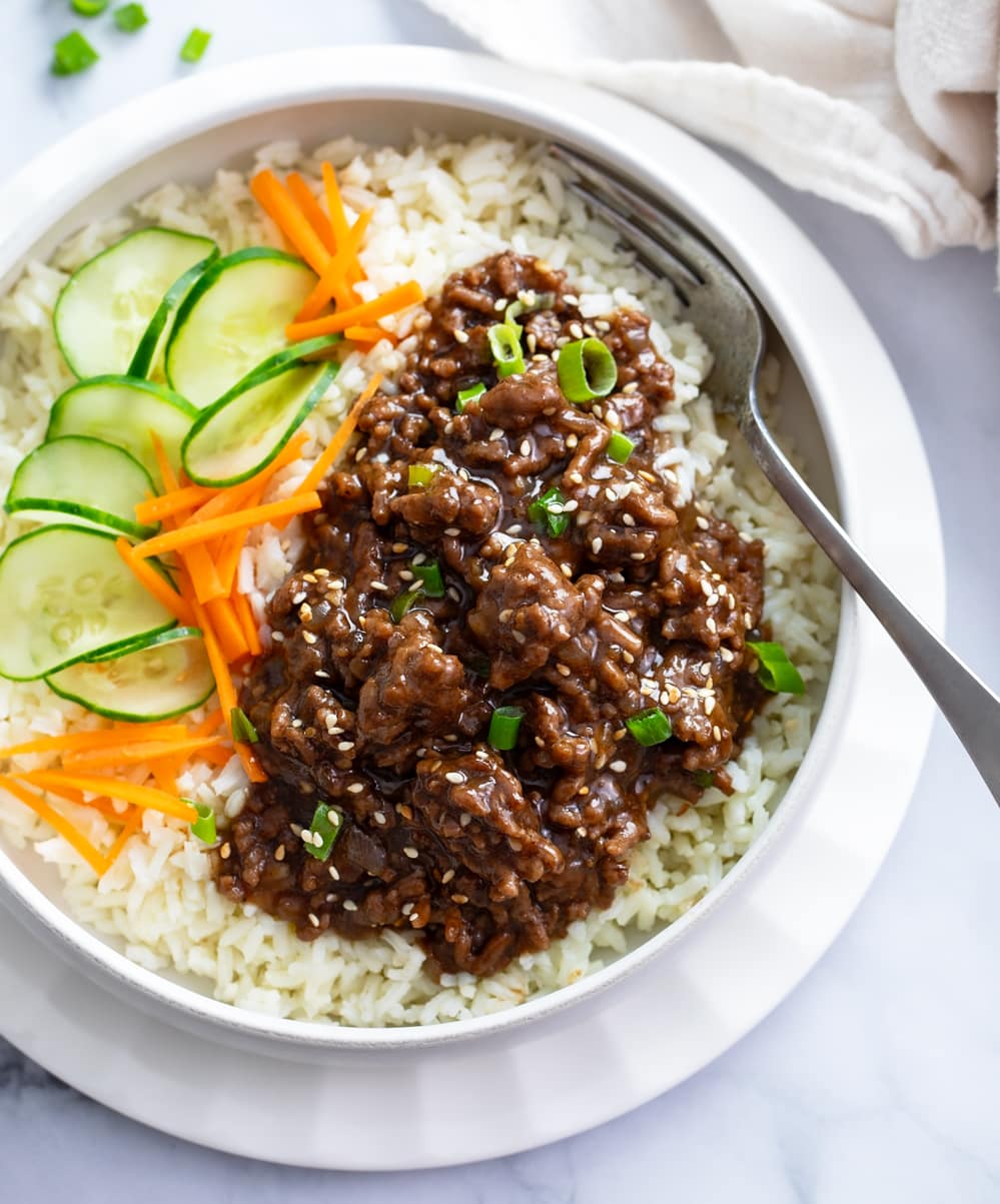 A white bowl of rice topped with Korean ground beef in sauce next to cucumbers and carrots.