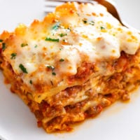 A square slice of cheesy Lasagna with layers of sauce and meat on a white plate.