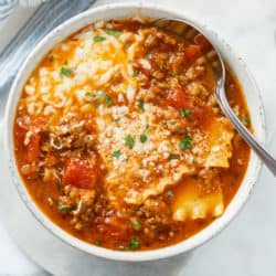 A white bowl filled with Lasagna Soup topped with cheese and a spoon on the side.