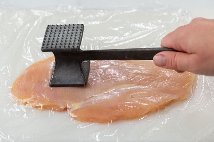 A hand holding a meat tenderizer and pounding a slice of chicken breast with saran wrap over it.