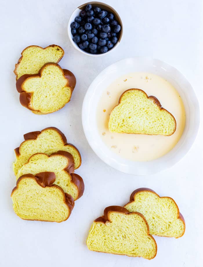 Challah bread slices being dipped into French toast custard on white table next to a bowl of blueberries.