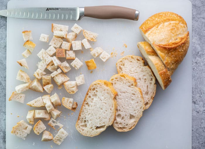 A loaf of Italian bread being cut into cubes on a white cutting board.