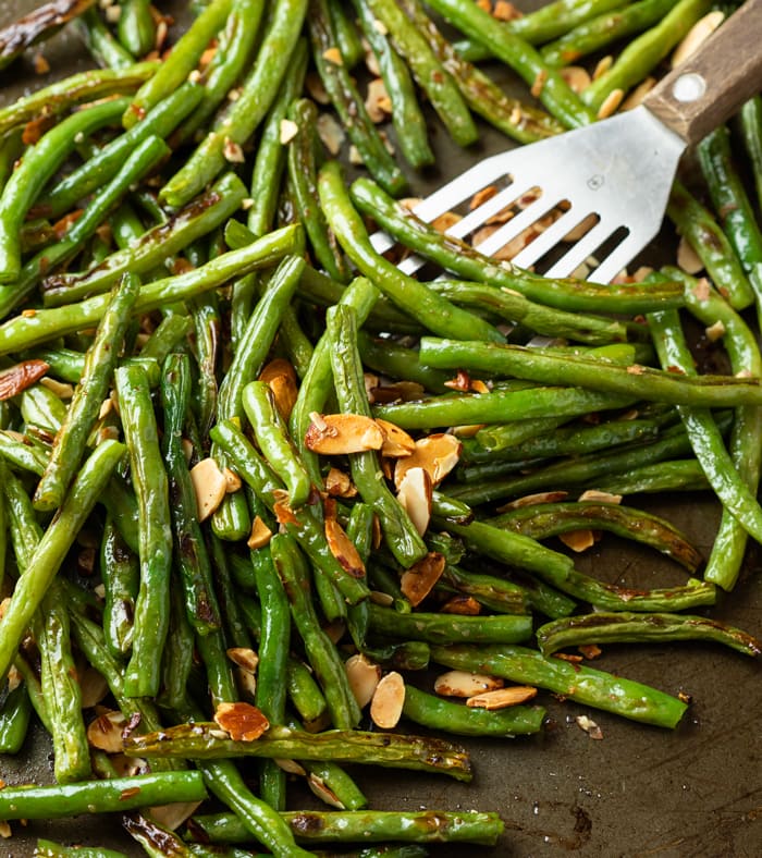 A dark roasting pant with a spatula scooping up roasted green beans topped with sliced almonds.