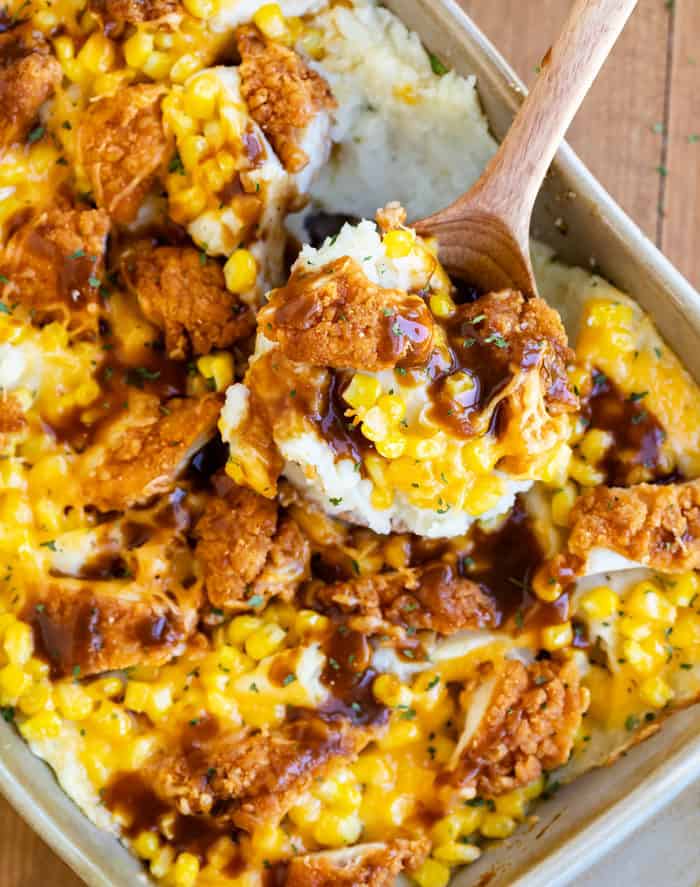 Casserole dish filled with mashed potatoes, corn, crispy chicken, cheese, and gravy with a wooden spoon scooping it out.