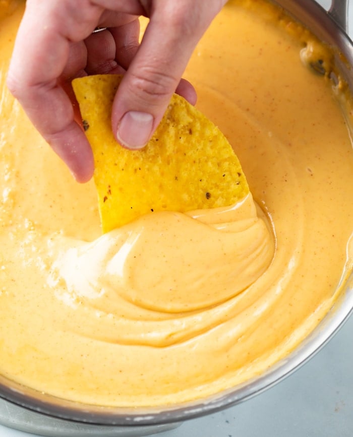 A hand dipping a tortilla chip into nacho cheese sauce.