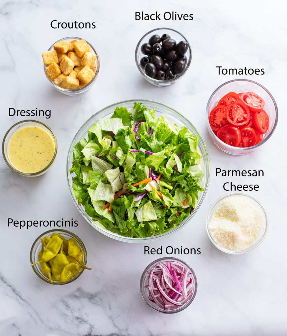 Labeled ingredients for making Olive Garden Salad on a white surface.