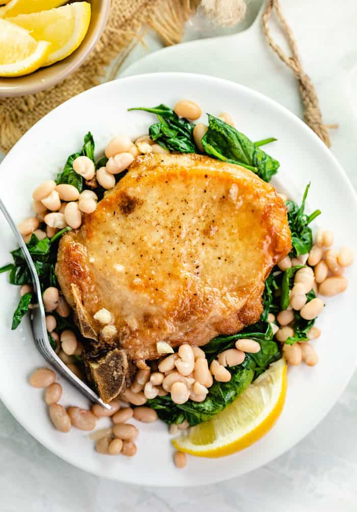 overhead shot of pan fried pork chops on a bed of sauteed spinach with white beans and a lemon wedge on a white plate. 