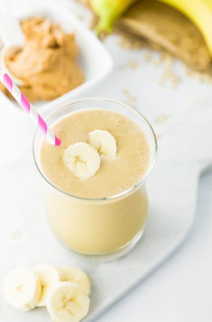 overhead image of a peanut butter banana smoothie in a glass with a scoop of peanut butter and bananas in the background. 