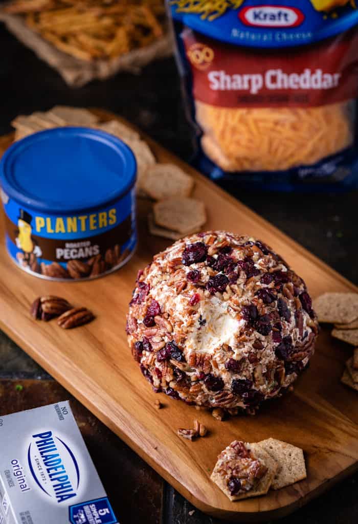 Wooden Cutting Board with Pineapple Cranberry Cheese Ball with planters pecans and grated cheese in background.