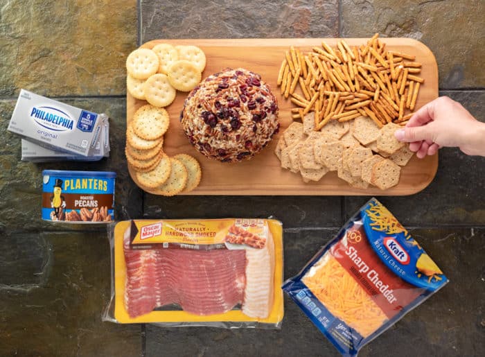 Wooden cutting board with cheese ball, crackers, pretzels, surrounding by a package of bacon, cheese, pecans, and cream cheese