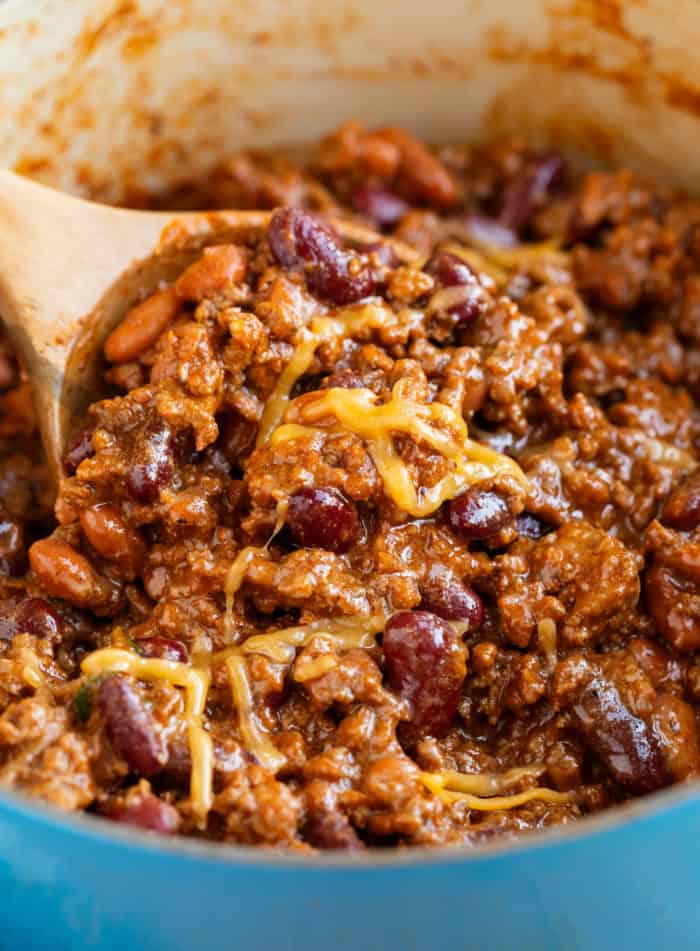 Close up view of a wooden spoon scooping chili out of a pot with melted cheese on top.