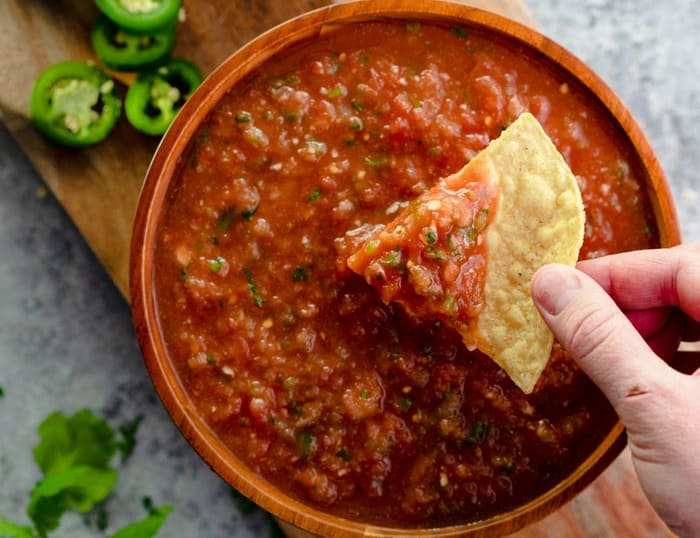 A bowl of salsa with a hand dipping a chip into it. Jalapenos are in the background.