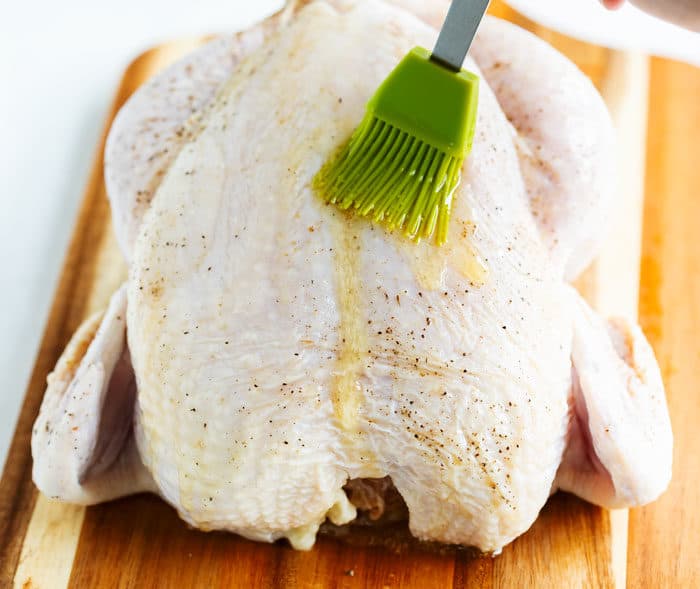A raw chicken on a wooden cutting board with a green pastry brush spreading melted butter over the top.