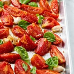 A rimmed baking sheet with roasted tomatoes and basil on top with salt and freshly cracked pepper.