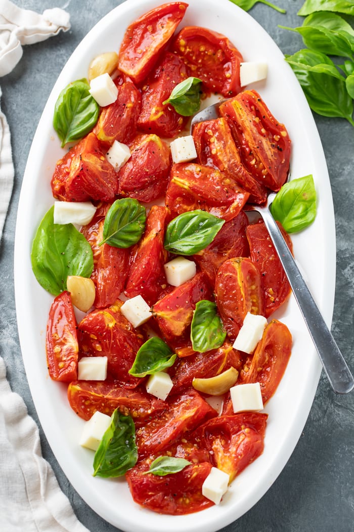 A white platter with Roasted tomatoes and basil with mozzarella, salt, and freshly cracked pepper.