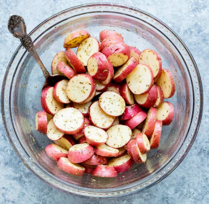Sliced and seasoned red potatoes in a large glass bowl with a spoon.