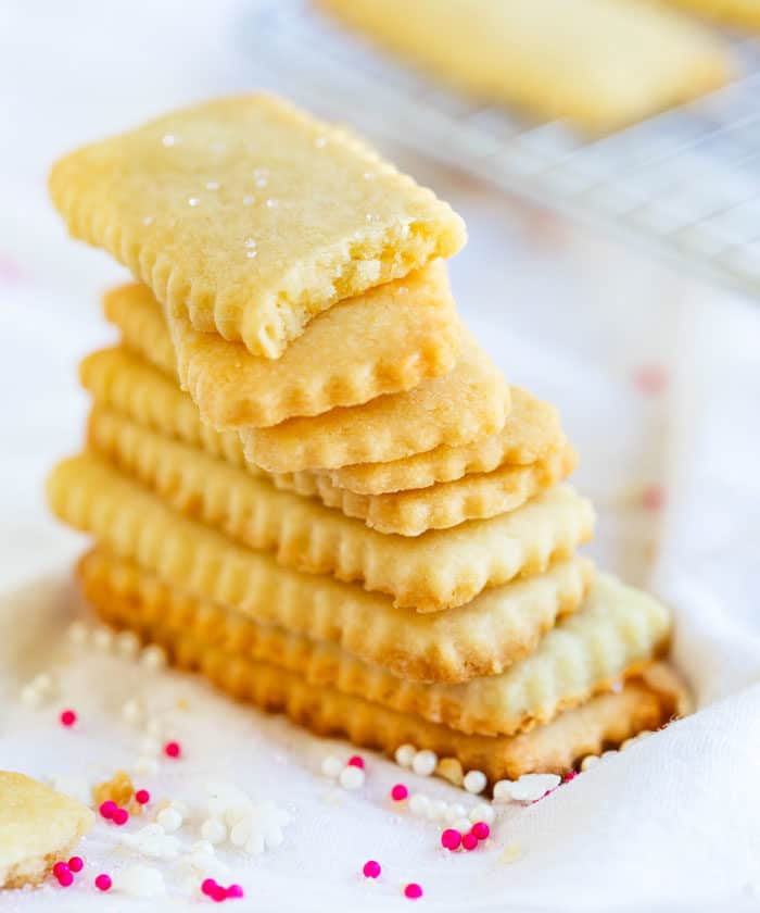 A tall stack of shortbread cookies on a white surface with sprinkles around it. Top cookies has a bite taken out of it.