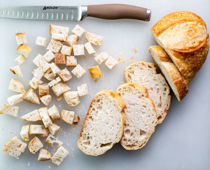 loaf of sourdough bread on a white cutting board with a knife that's partially cubed and sliced for homemade croutons.