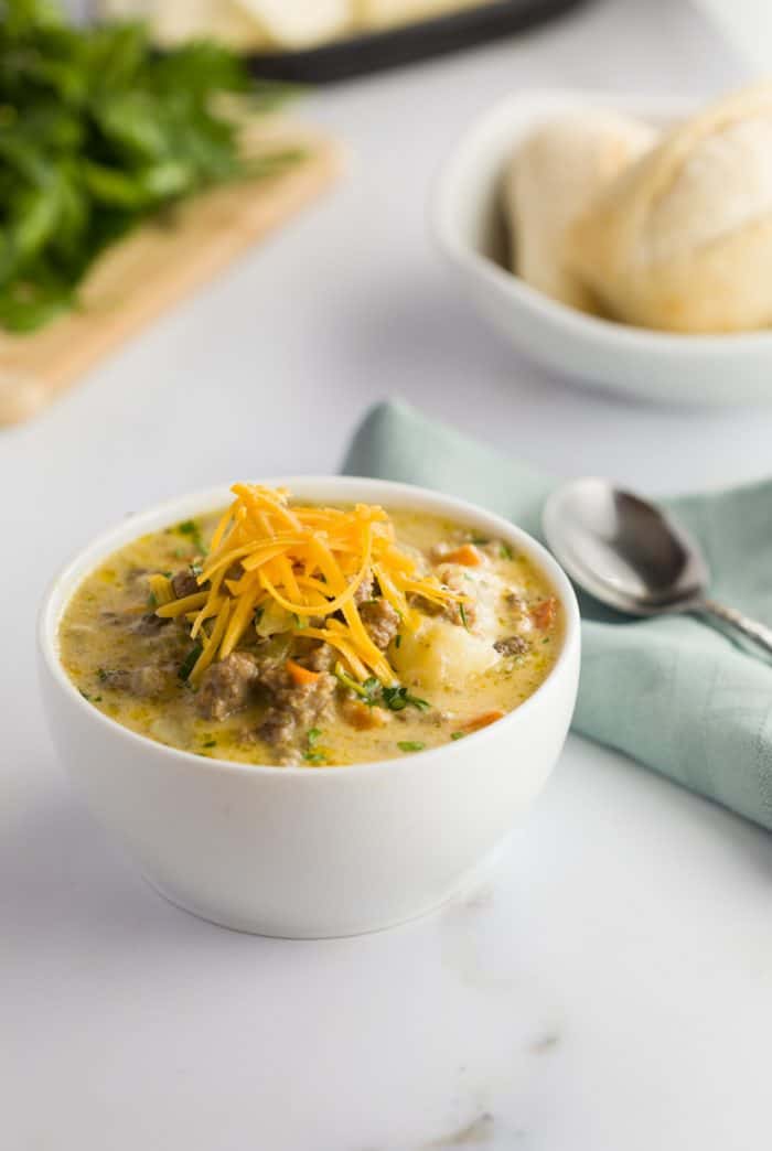 Slow Cooker Cheeseburger Soup in a white bowl on a white countertop with rolls in the background.