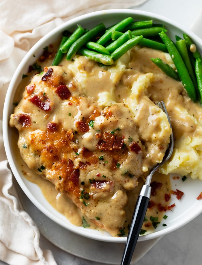 A white plate with Smothered Chicken next to mashed potatoes with gravy and green beans.