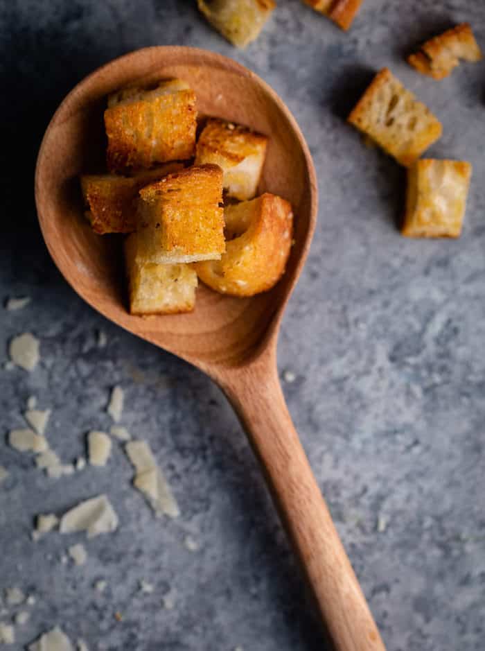 wooden spoon holding homemade croutons on a shadowy surface sprinkled with grated Parmesan. 