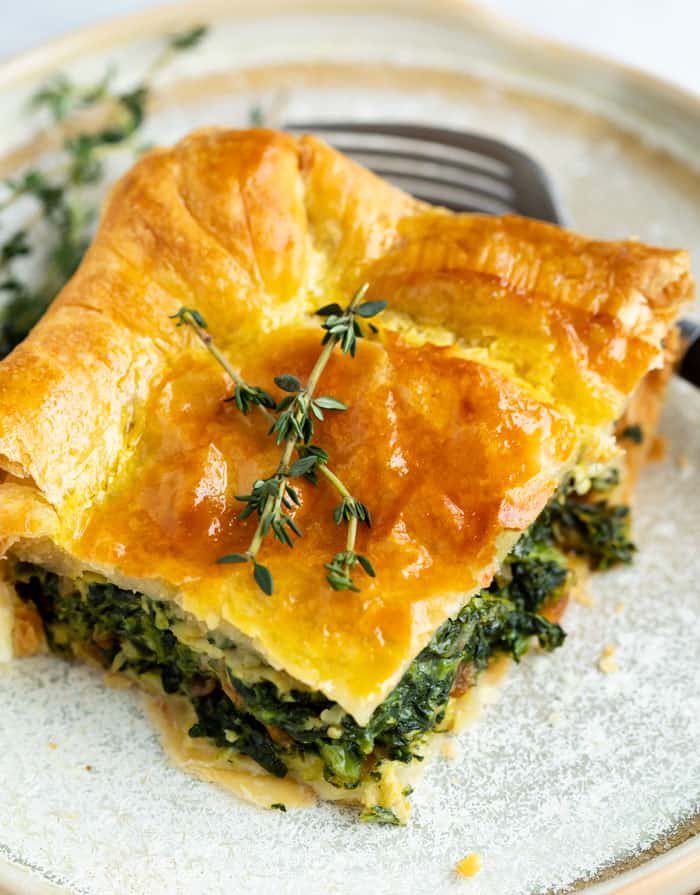 A slice of spinach pie with a golden brown puff pastry topping garnished with fresh thyme on a plate.