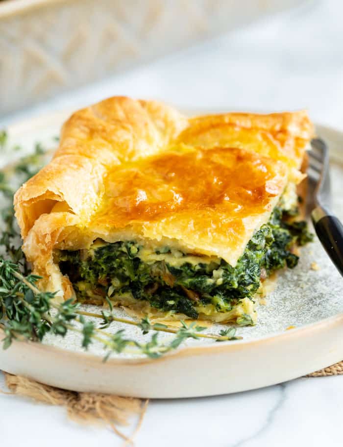 A slice of spinach pie on a plate with fresh thyme next to it and a fork on the side.
