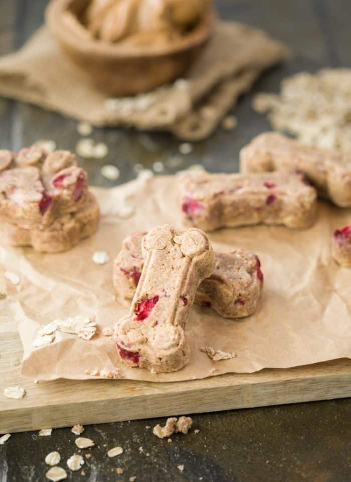 homemade dog treats in the shape of a dog bone sitting on crinkled brown paper on a cutting board with oats.