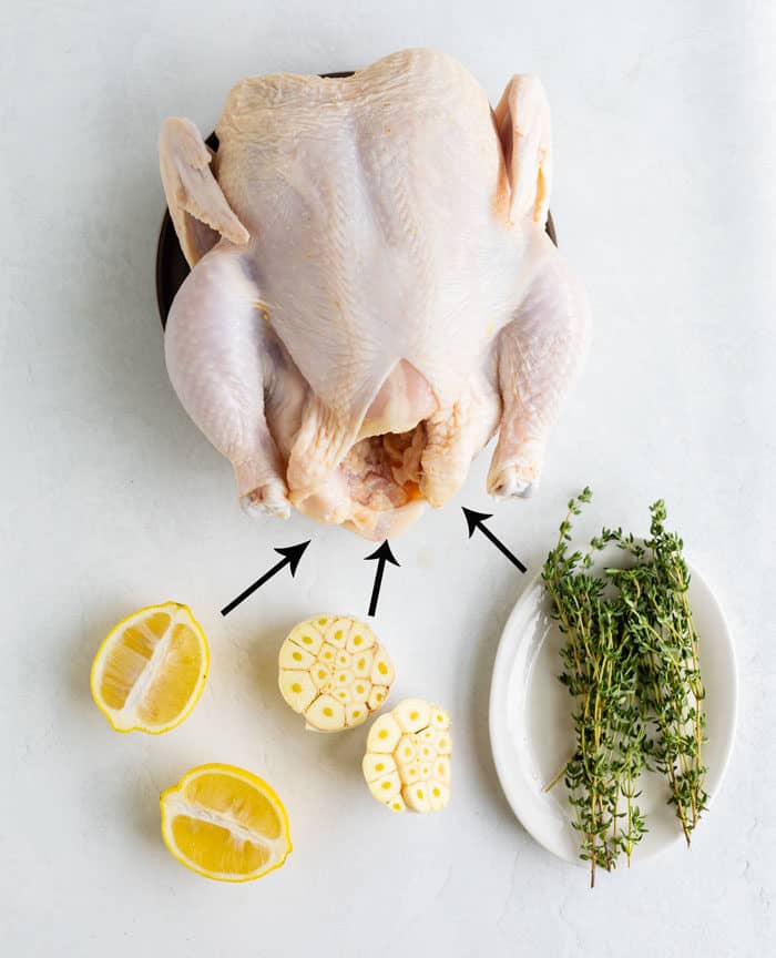 A raw chicken on a white surface with thyme, lemon, and garlic for stuffing the chicken with.