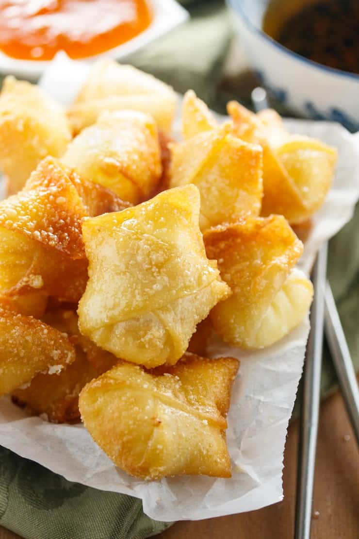 A pile of fried wontons with cream cheese filling and sweet and sour sauce in the background.
