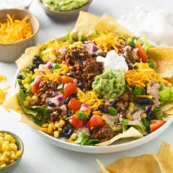 A white plate filled with Taco Salad topped with ground beef, avocado, sour cream, tortilla chips, and vegetables.