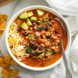 A white bowl filled with taco soup garnished with diced avocado and shredded cheese.