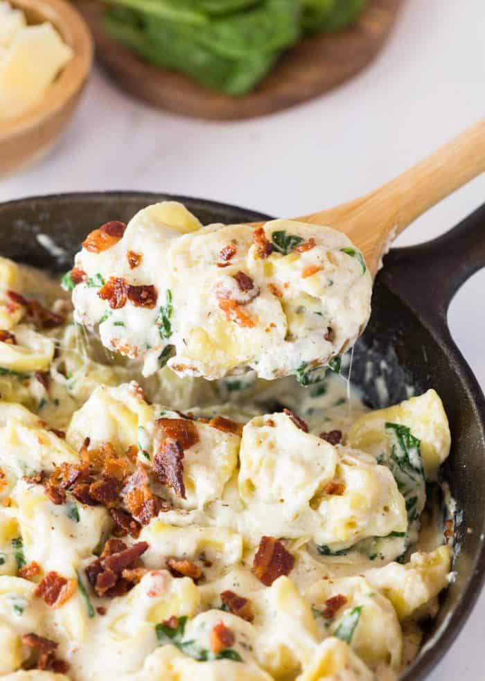 A cast iron skillet filled with tortellini in a cream sauce topped with bacon and a wooden spoon scooping some out.