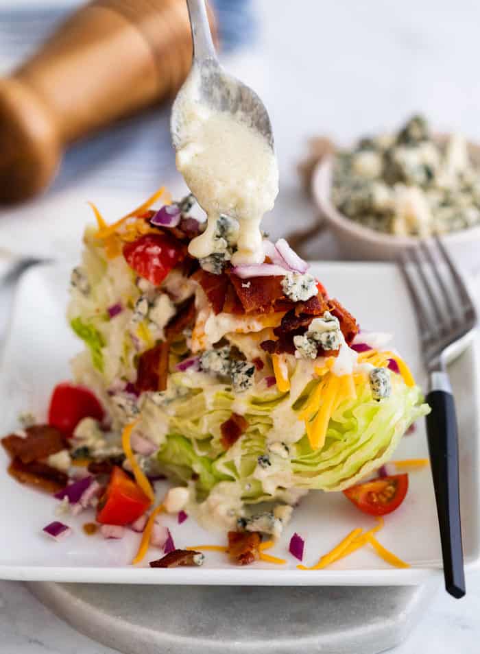 a spoon drizzling blue cheese on top of a wedge salad on a white plate.
