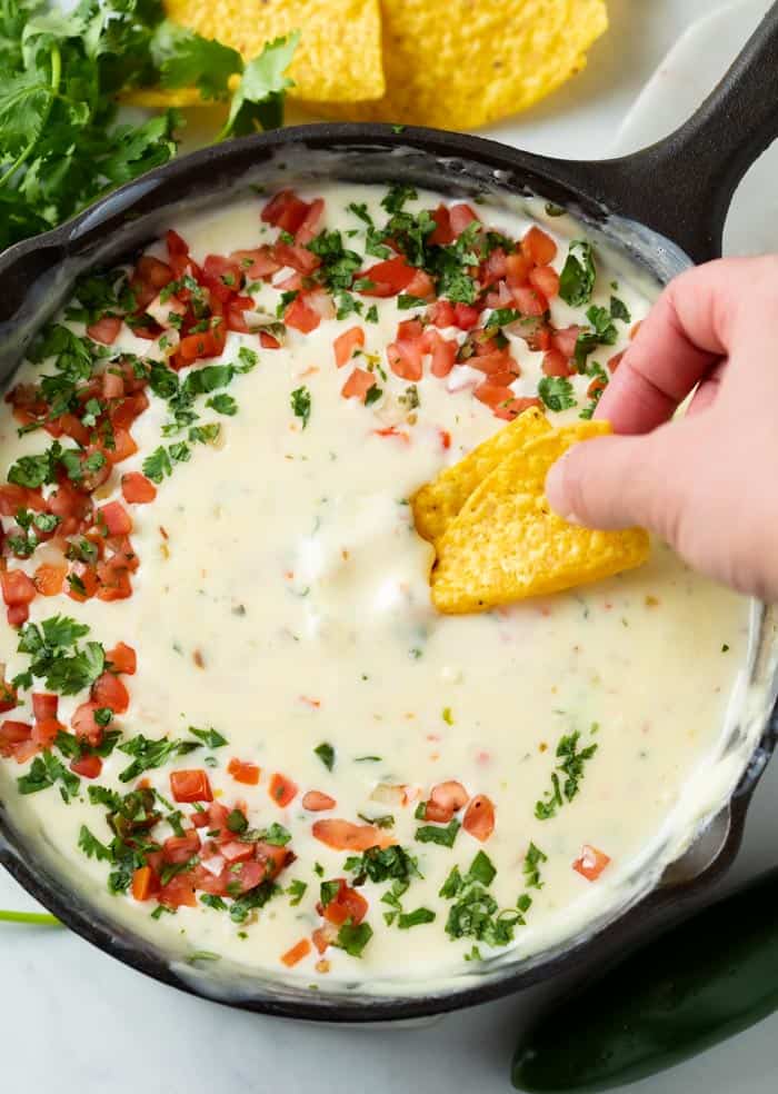 A hand dipping a tortilla chip into a skillet of White Queso Dip topped with Pico De Gallo and Cilantro