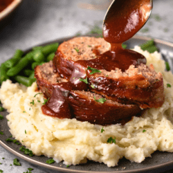 A pile of mashed potatoes with The Pioneer Woman Meatloaf with a spoon drizzling glaze on top.